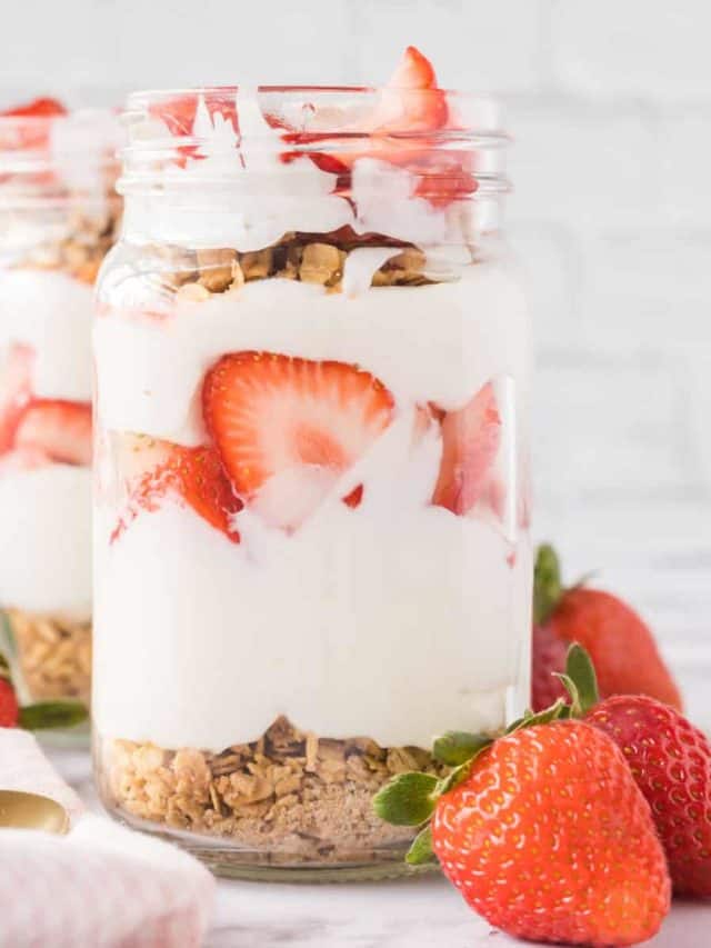 Two jars filled with strawberry breakfast parfaits set on a marble table with whole strawberrys set at the base of the jars and a tea towel and spoons set to the side.