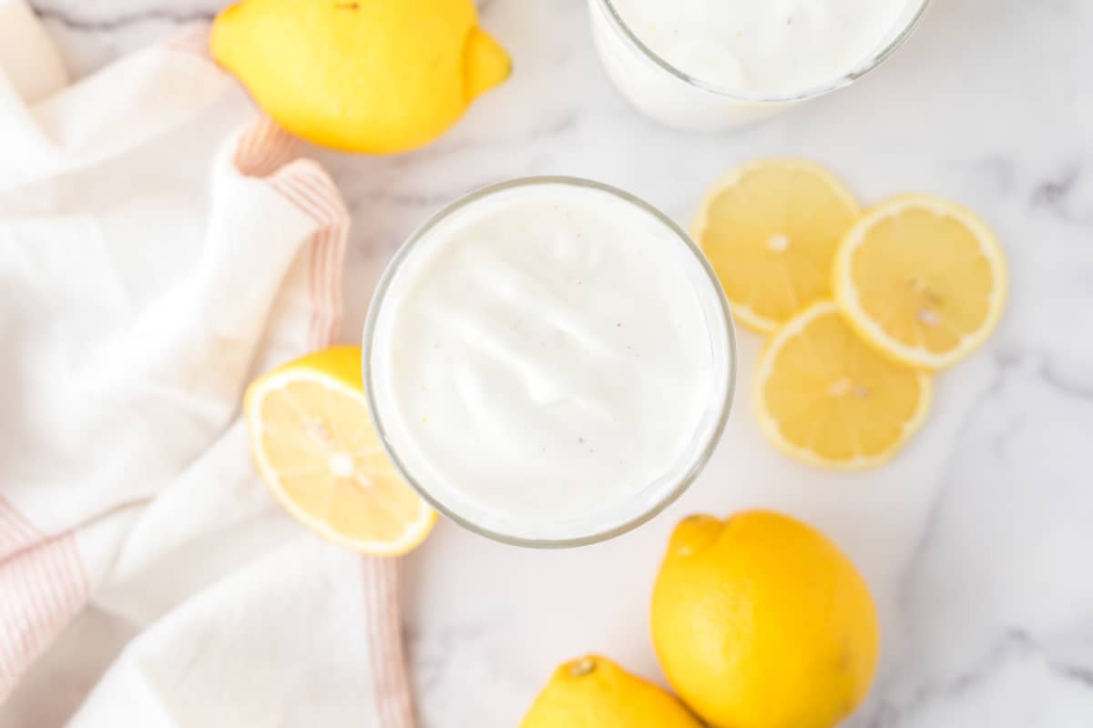 An overhead shot of a glass of creamy frosted lemonade on a marble table with lemons scattered about.