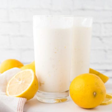 Two glasses filled with frosted lemonade with lemons surrounding glass on the table.