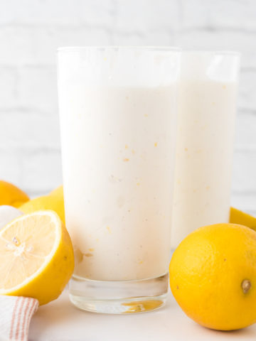 Two glasses filled with frosted lemonade with lemons surrounding glass on the table.
