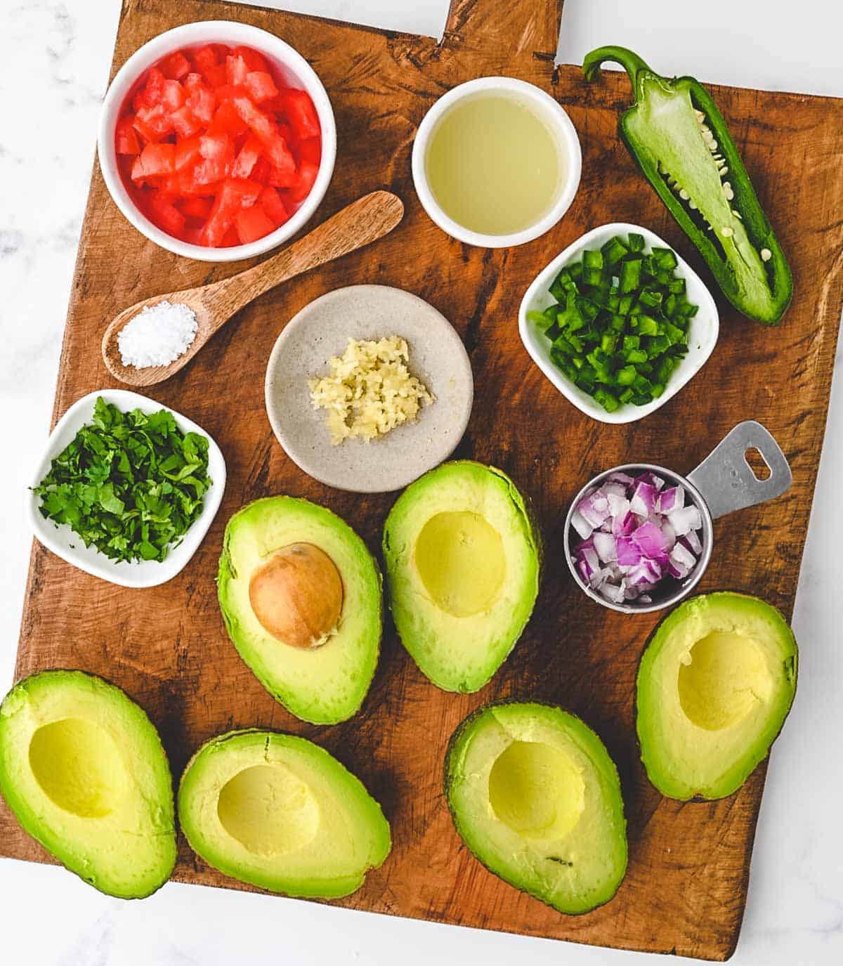 A cutting board with avocado halves, diced tomatoes, lime juice, diced jalapenos, minced garlic, chopped cilantro, diced red onion, and coarse sea salt.