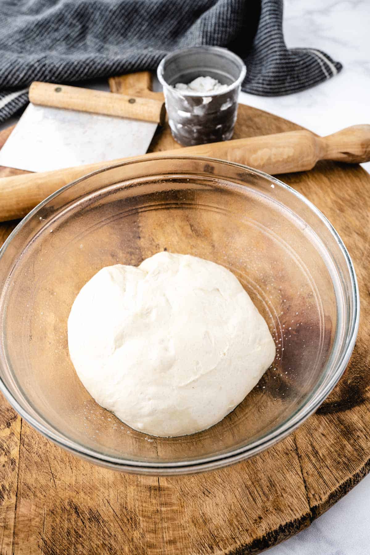 A ball of pizza dough in an oiled glass bowl set on a wooden tray.