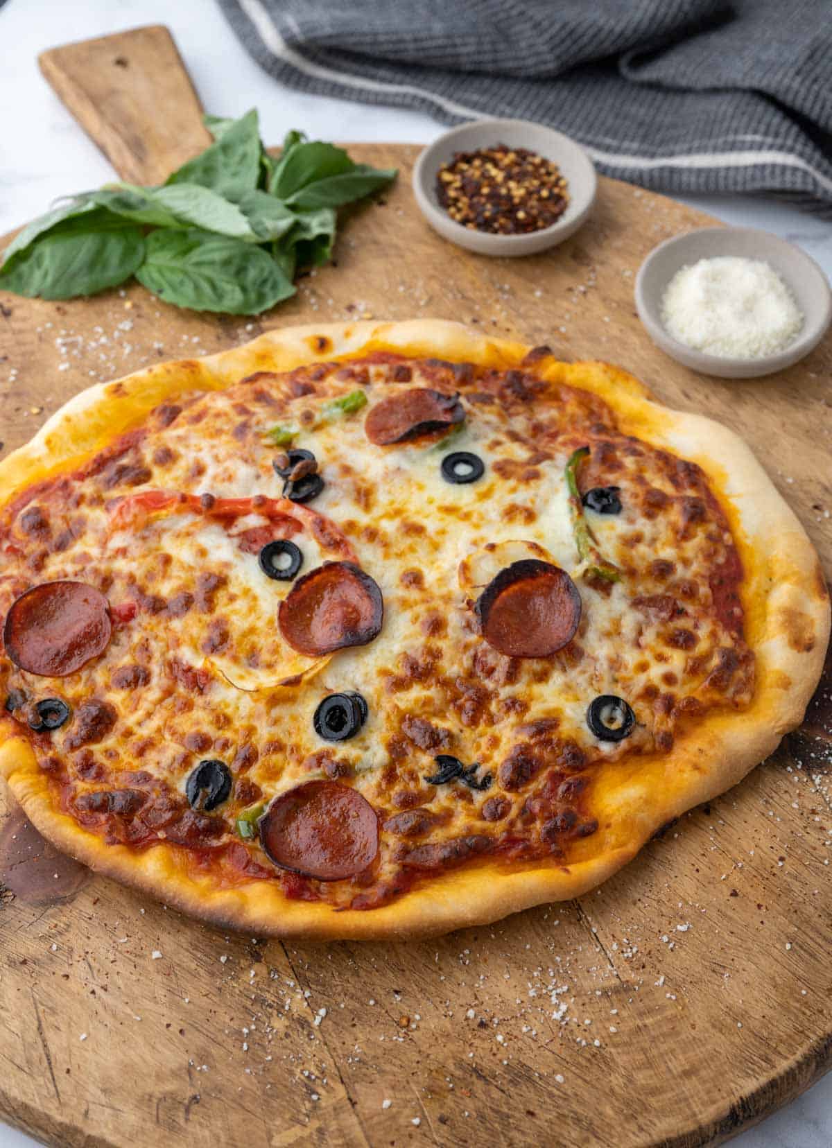 A homemade pizza topped with pepperoni, mozzarella, cheese, peppers, and olives set on a wooden board garnished with basil, red pepper flakes, and parmesan cheese.