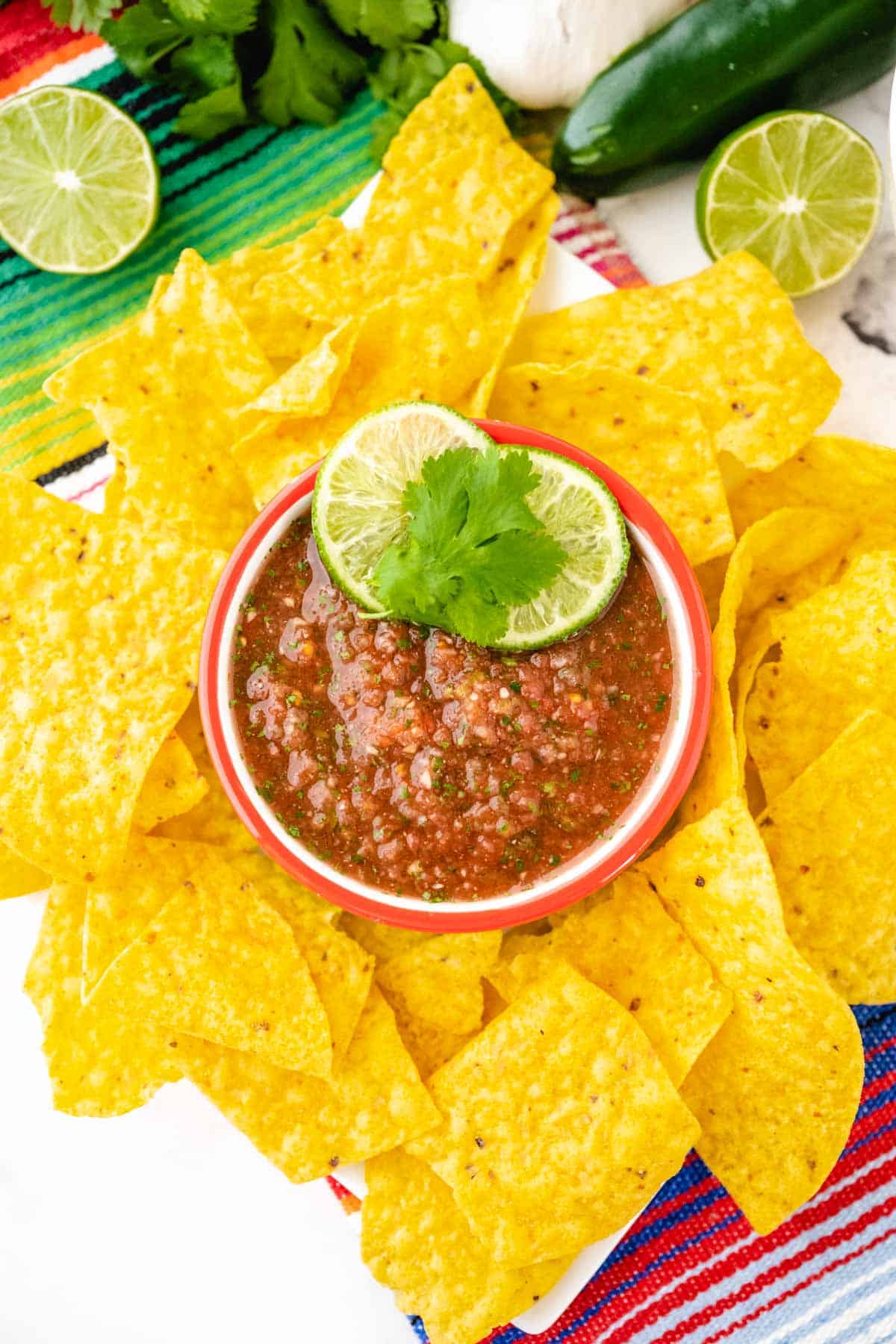 An overhead image of a bowl of restaurant style salsa on a Mexican themed cloth with cilantro, lime, garlic, an jalapenos in the background.