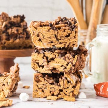 S'mores cereal bars stacked three high on a marble counter with marshmallows and golden graham cereal scattered on the table.