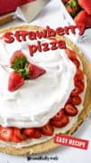 A whole strawberry pizza on a white wooden table with a carton of berries in the background.