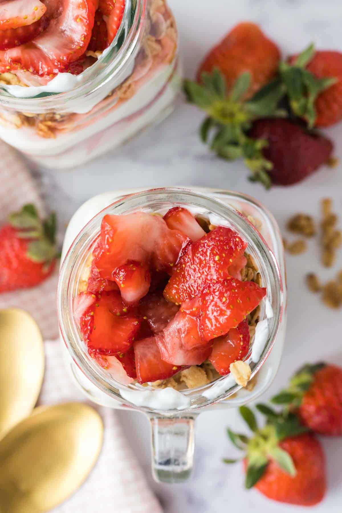 An overhead image of the top of a strawberry, yogurts, and granola filled mug on a tablescape of berries, granola, and spoons to the side.