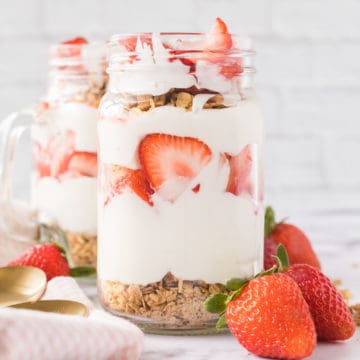 Two jars filled with strawberry breakfast parfaits set on a marble table with whole strawberrys set at the base of the jars and a tea towel and spoons set to the side.