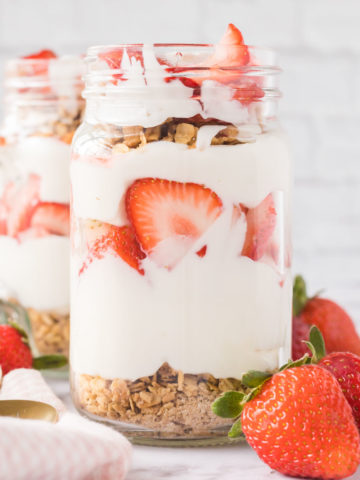 Two jars filled with strawberry breakfast parfaits set on a marble table with whole strawberrys set at the base of the jars and a tea towel and spoons set to the side.