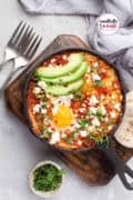 A cast iron pan with tomato sauce topped with poached eggs garnished with avocado and feta.