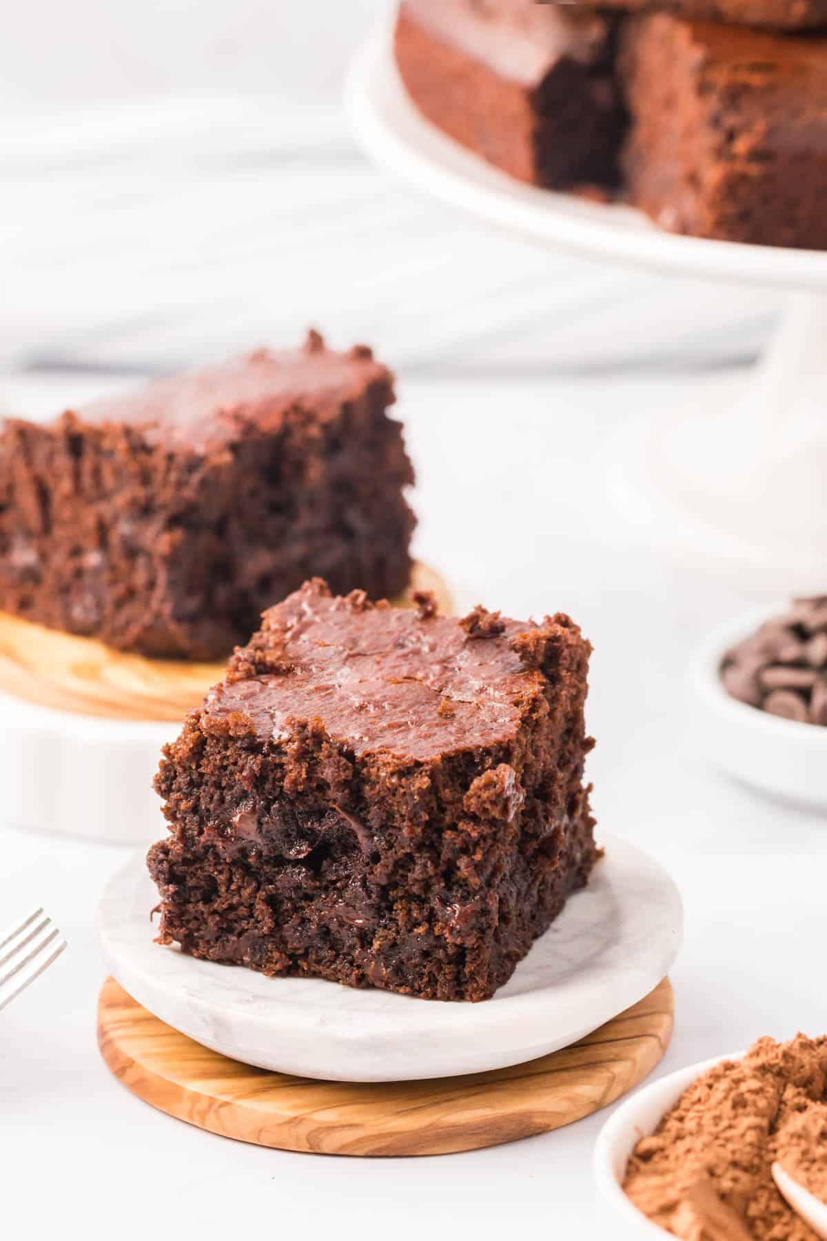 A cake-like brownie slice on a white plate with another slice on a wooden plate behind it, with a cake pedestal filled with brownies in the background.