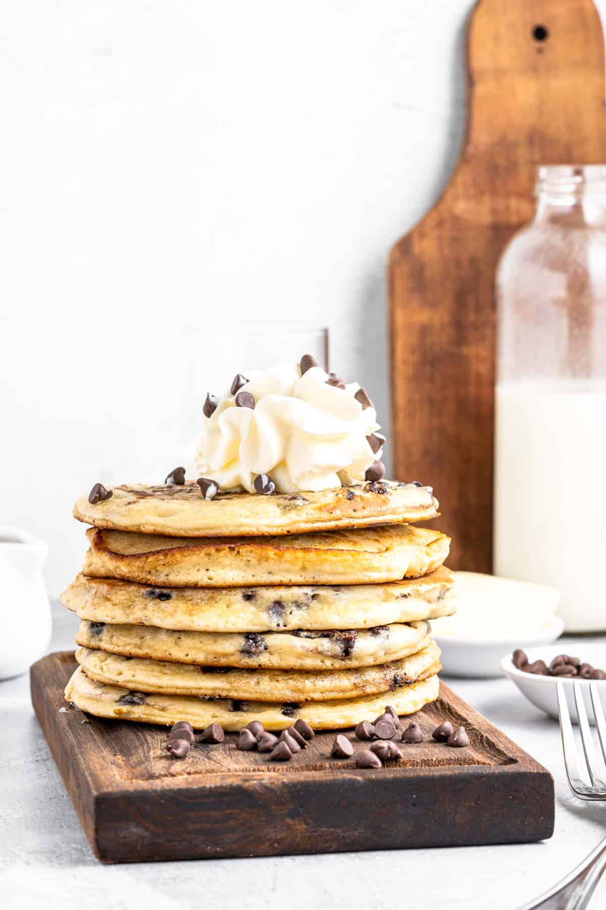 A stack of chocolate chip pancakes topped with whipped cream and chocolate chips on a wooden tray.