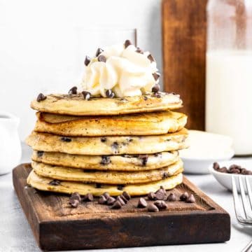 A stack of chocolate chip pancakes topped with whipped cream and chocolate chips on a wooden tray.