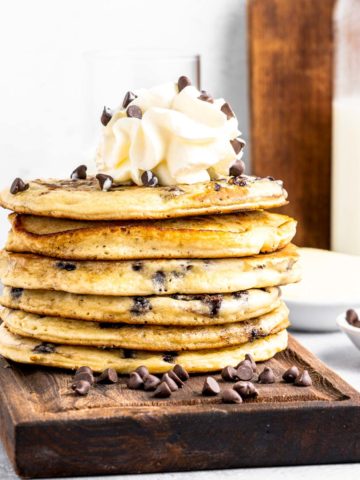 A stack of chocolate chip pancakes topped with whipped cream and chocolate chips on a wooden tray.