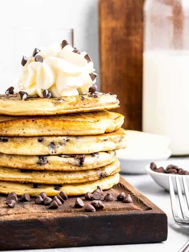 A stack of chocolate chip pancakes topped with whipped cream and chocolate chips on a wooden tray.