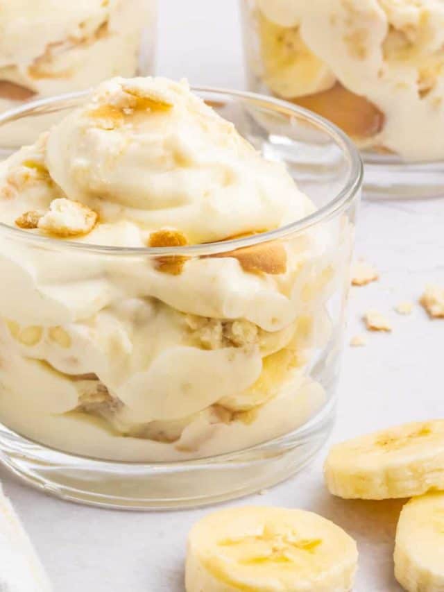 Three desert cups filled with Magnolia Bakery Banana Pudding set on a table with sliced bananas and a white tea towel set in the front.