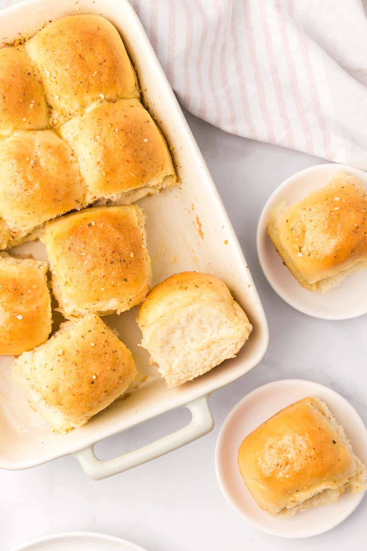 A white baking dish filled with homemade dinner rolls on a white table with two rolls on bread plates off to the side.