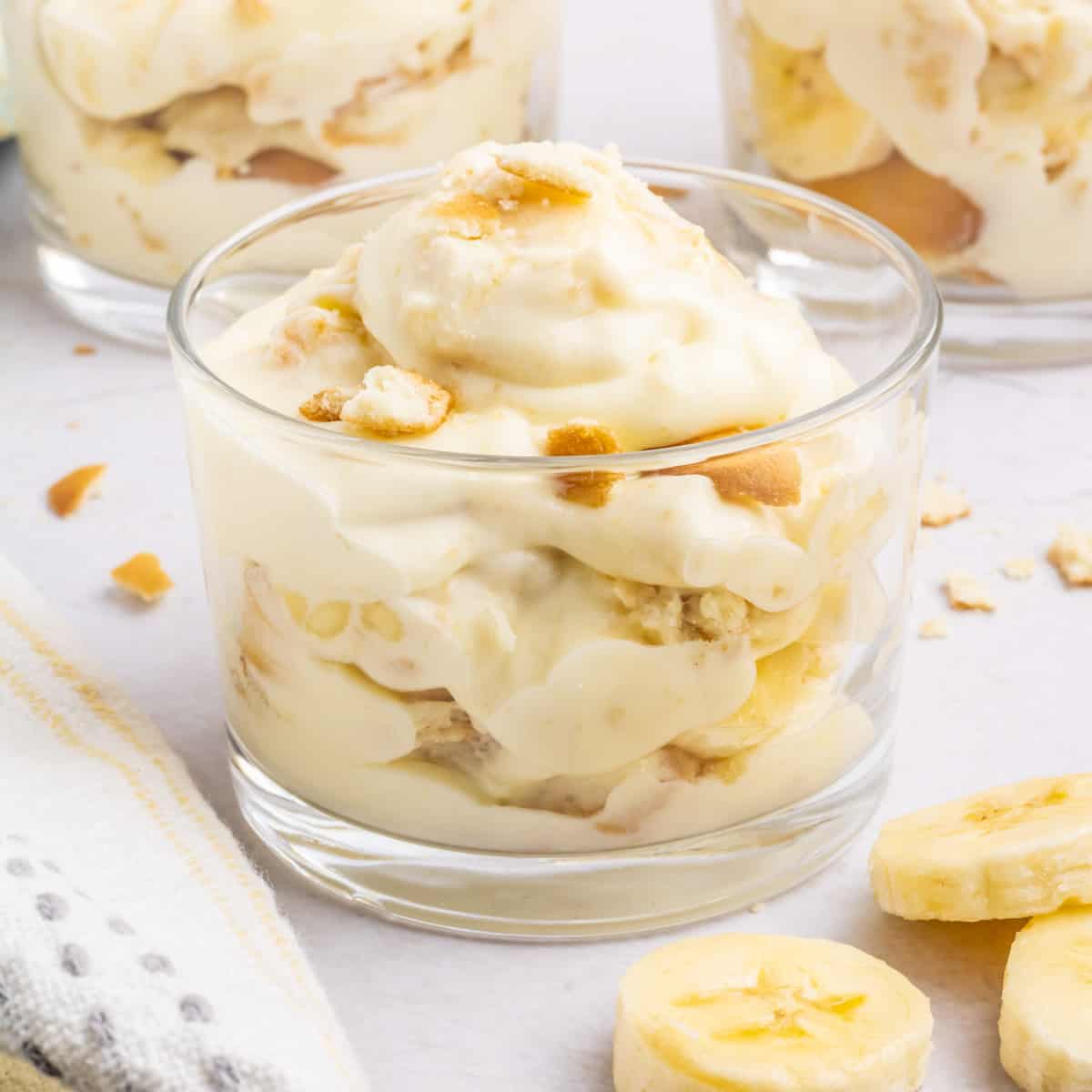 Three desert cups filled with Magnolia Bakery Banana Pudding set on a table with sliced bananas and a white tea towel set in the front.