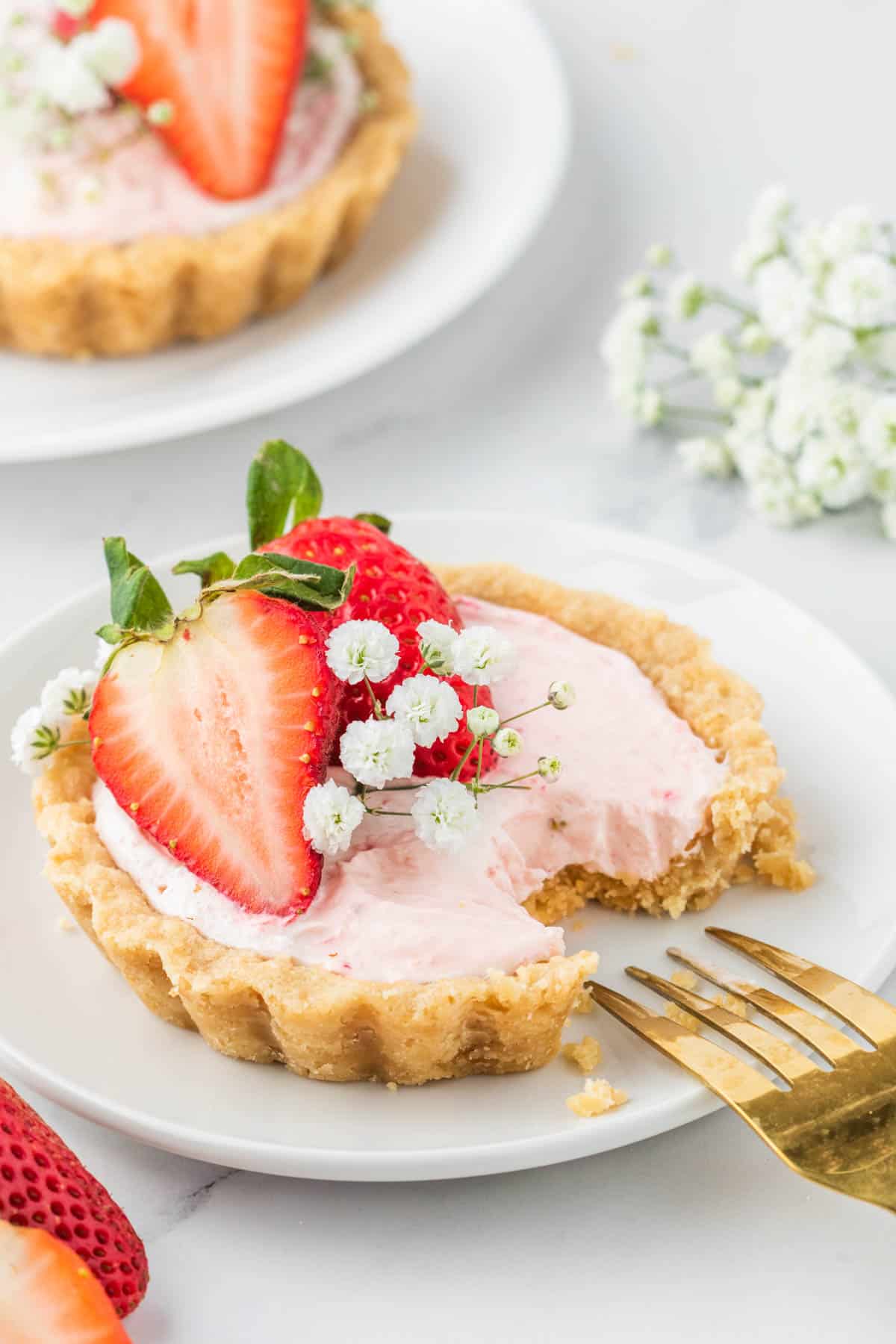 A mini strawberry tart set on a white plate with a bite removed with a gold fork.