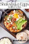 Shakshuka eggs topped with sliced avocado and feta cheese in a skillet.