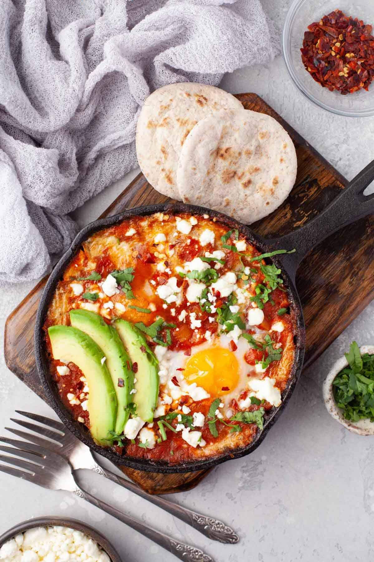 Shakshuka in a cast iron skillet set on a wooden board with pita and spices.