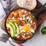 Shakshuka topped with feta, avocado, and cilantro on a wooden tray.