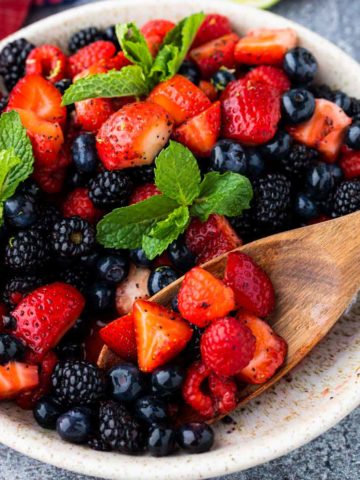 Berry Fruit Salad tossed in a honey lime dressing in a rustic bowl with some of the berries being scooped out with a wooden spoon.