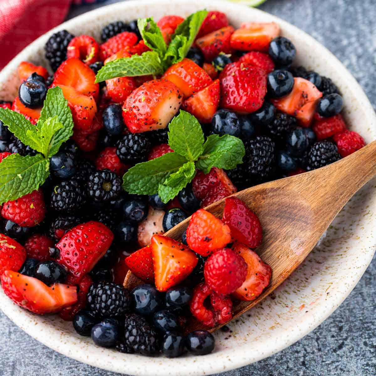 Berry Fruit Salad tossed in a honey lime dressing in a rustic bowl with some of the berries being scooped out with a wooden spoon.