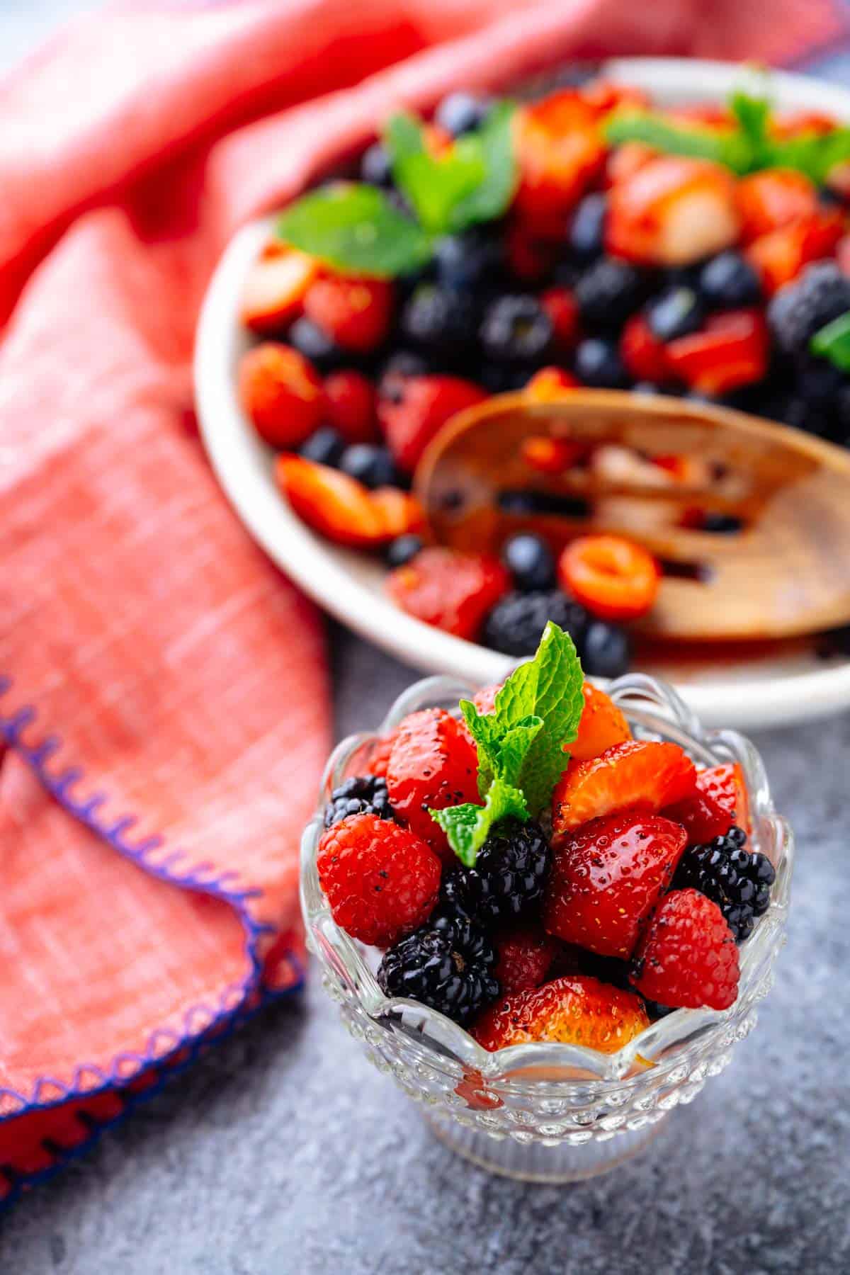 A glass dessert cup filled with berry fruit salad with the serving bowl of fruit salad in the background.
