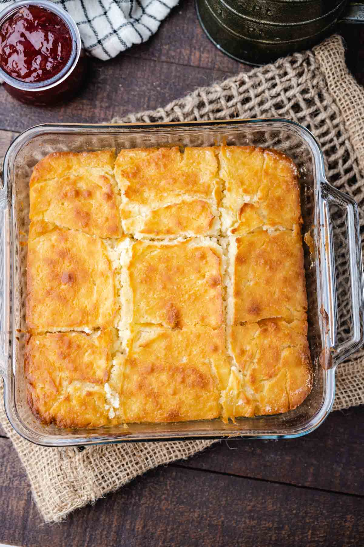 An overhead view of a glass baking dish filled with butter swim biscuits on a wooden table with strawberry jelly in the background.