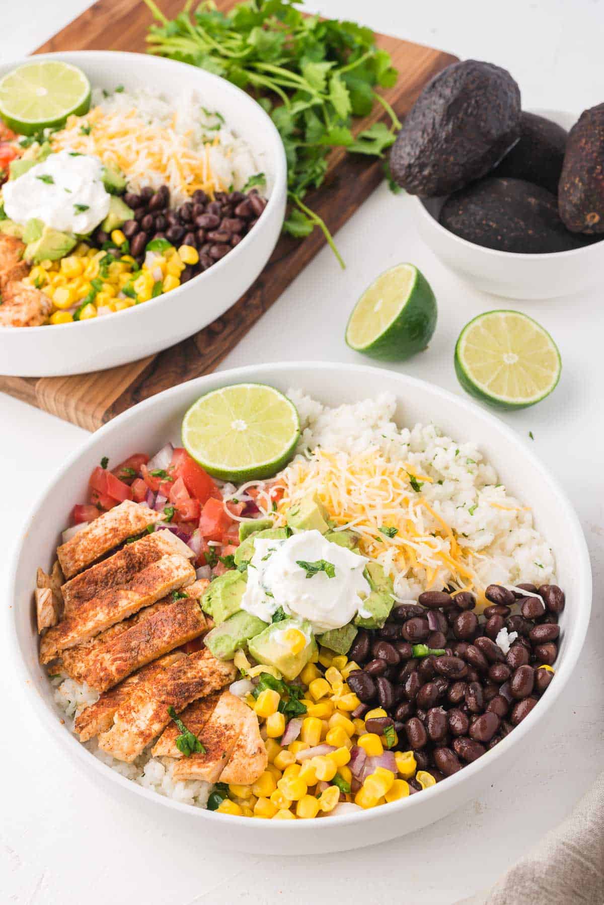 A chicken burrito bowl on a white table with another bowl in the background styled with a bowl of whole avocados and a lime sliced in half.