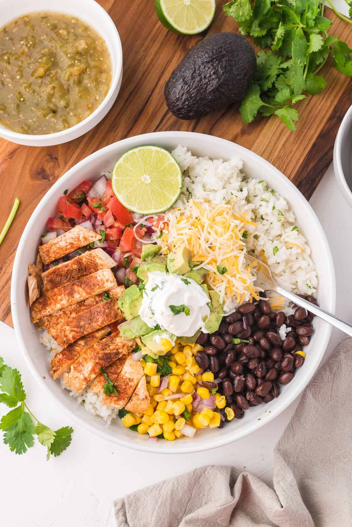 A bowl of chipotle copycat chicken burrito bowl on a table with a bowl of salsa verde and avocados.