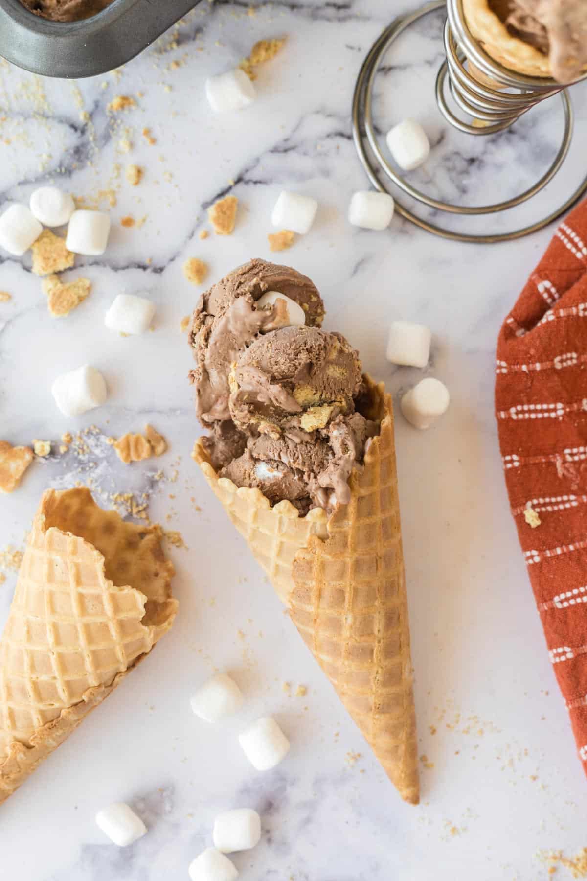 A waffle cone filled with chocolate s'mores ice cream on a marble table with scatter mini marshmallows and graham cracker crumbs on the table.