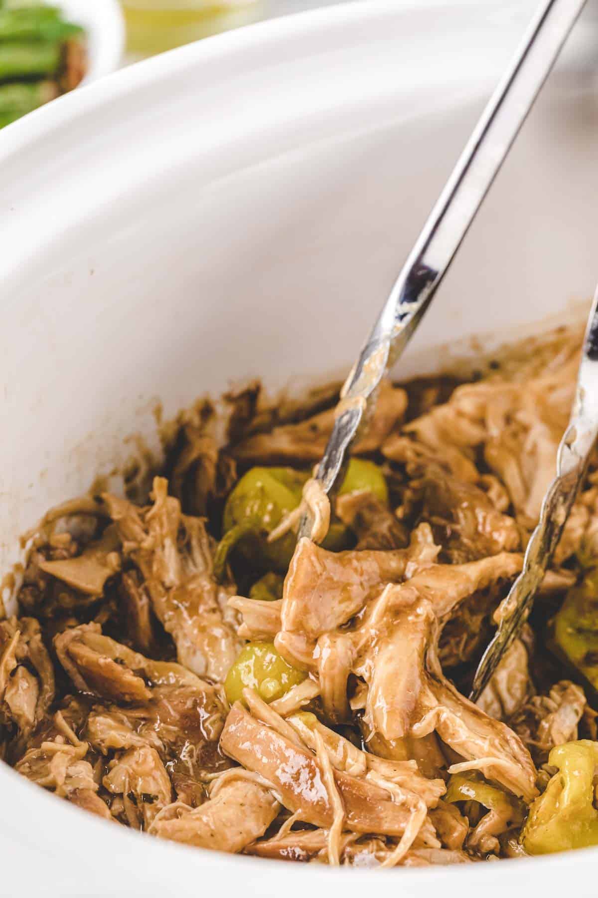 Tongs stirring chicken in a white slow cooker crock.