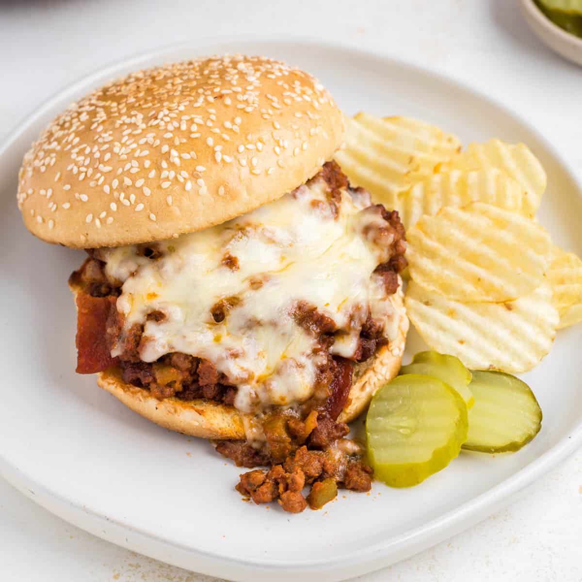 A pizza sloppy joe with melted mozzarella on a toasted sesame seed bun plated with pickles and chips.