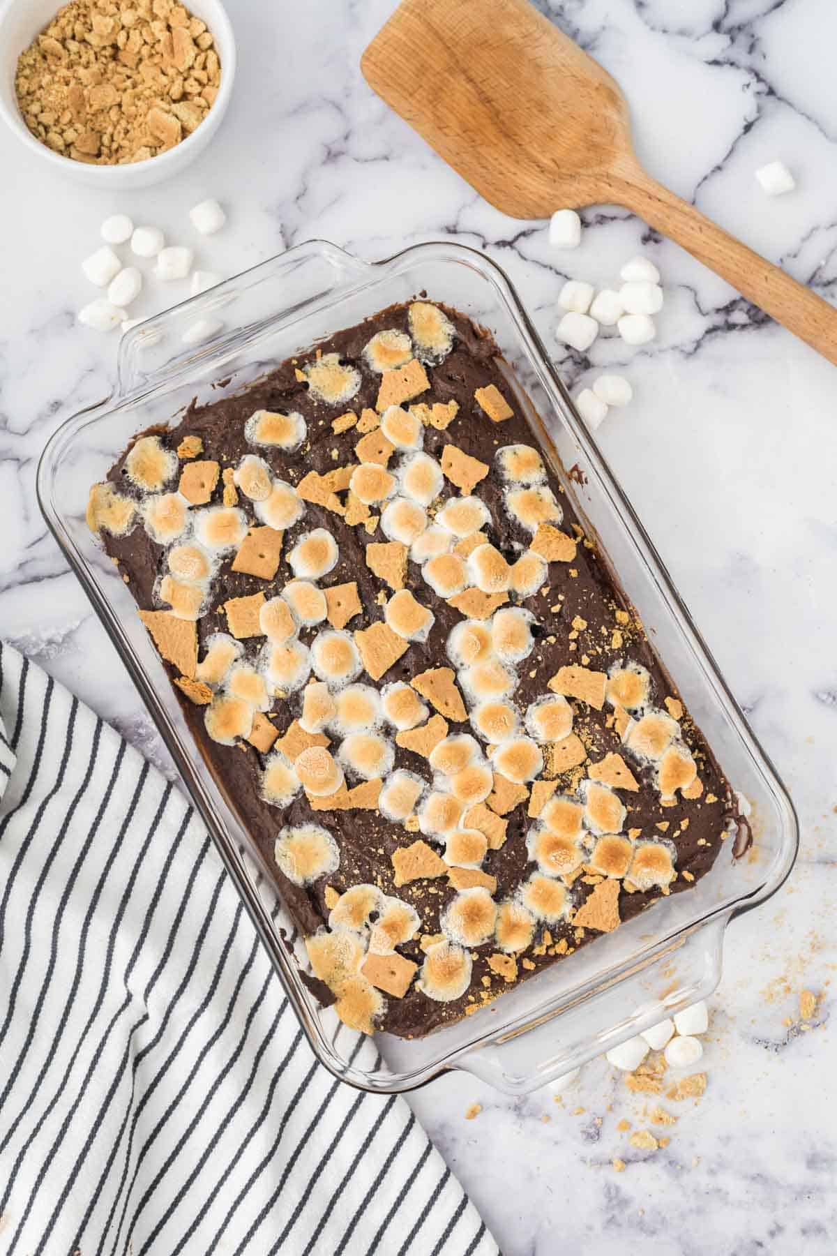 A glass pan filled with s'mores brownies topped with toasted mini marshmallows and graham cracker chunks.