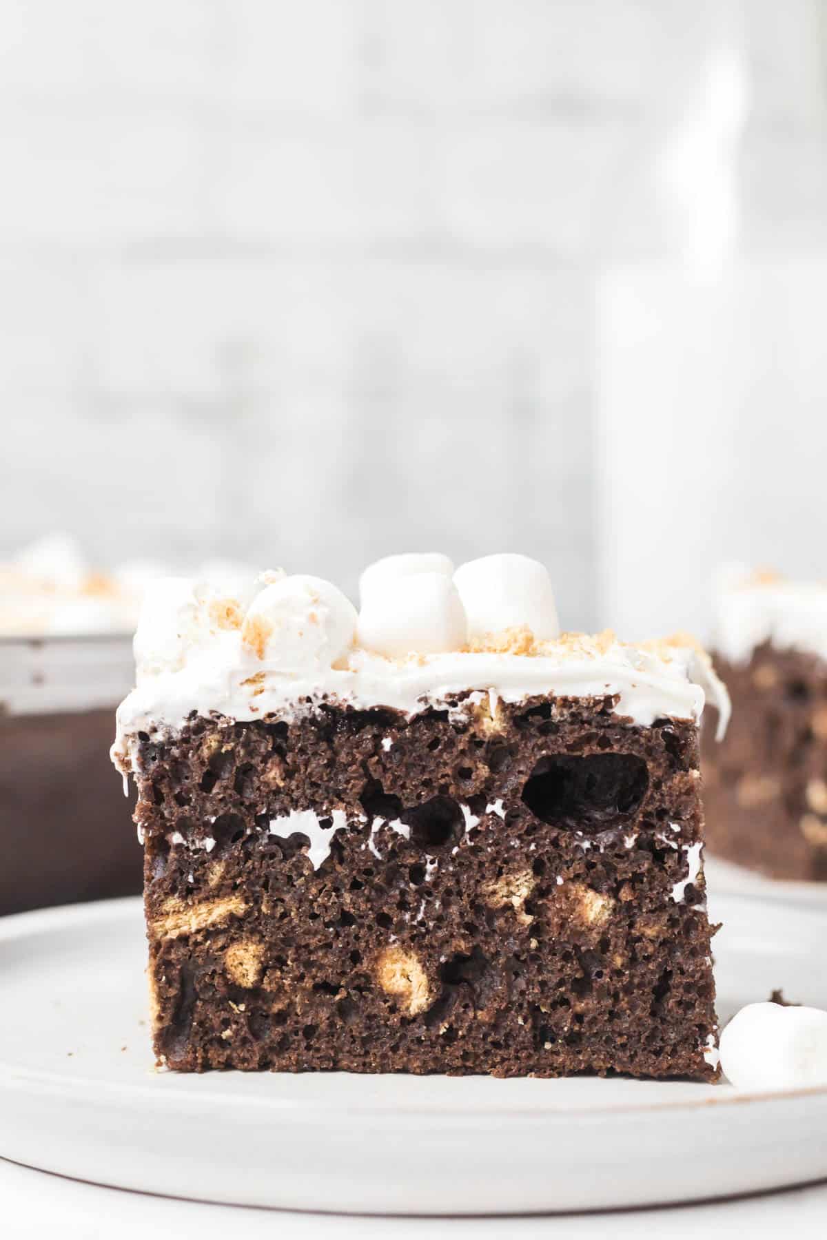 A chocolate easy s'mores cake recipe with crushed graham crackers topped with marshmallow frosting on a white plate.