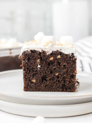 A slice of S'mores cake on a white plate with the can pan, a carafe of milk, and a tea towel in the background.