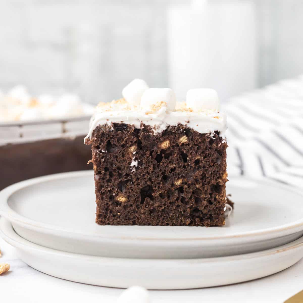 A slice of S'mores cake on a white plate with the can pan, a carafe of milk, and a tea towel in the background.