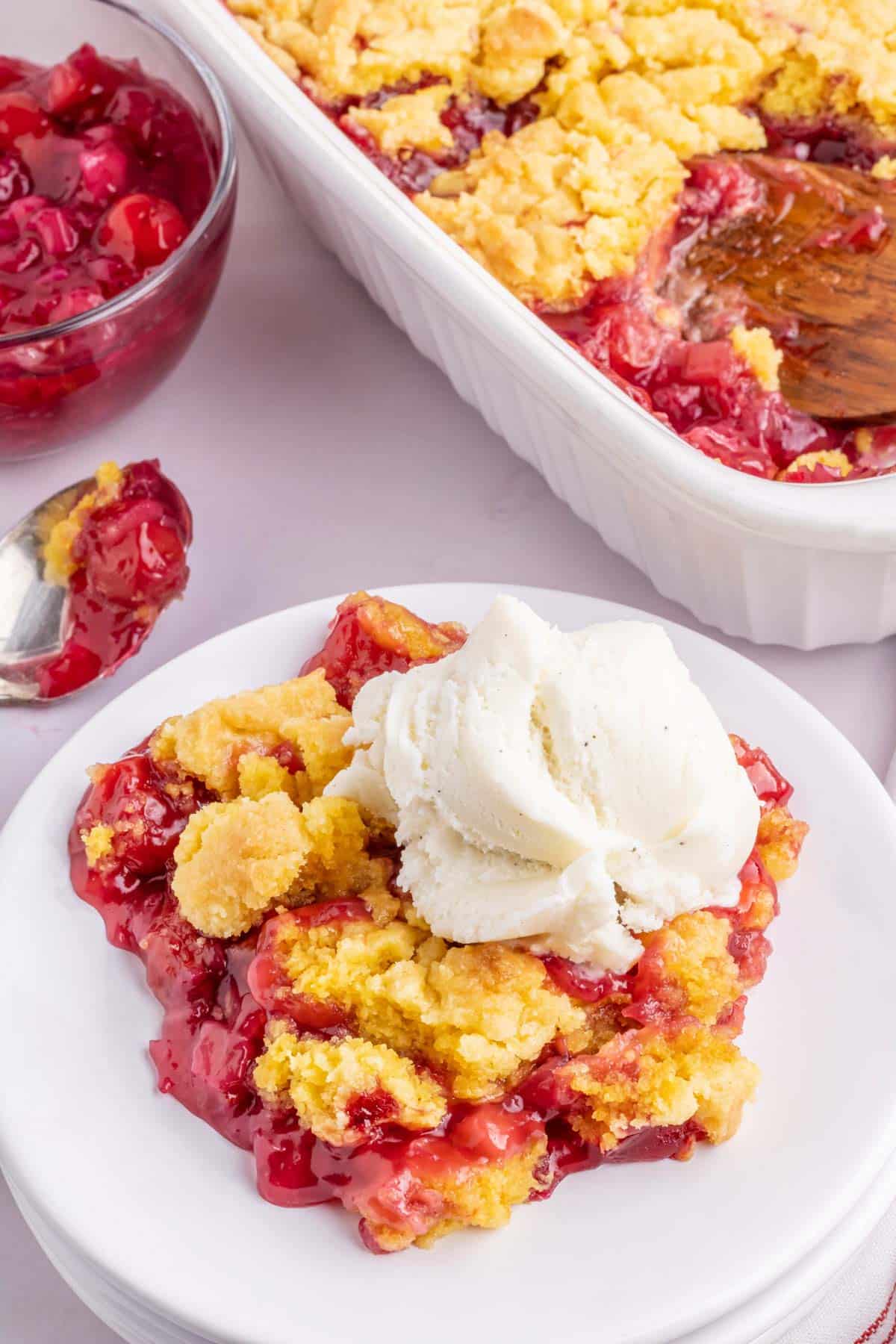 A white plate filled with a serving of cherry pineapple dump cake topped with a scoop of vanilla ice cream.