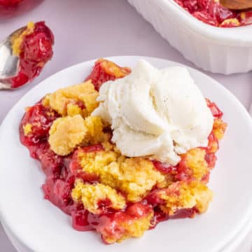 A white plate filled with a serving of cherry pineapple dump cake topped with a scoop of vanilla ice cream.