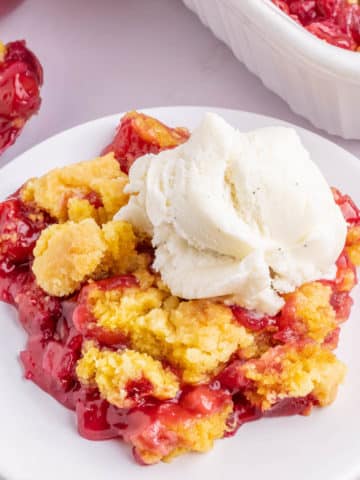 A white plate filled with a serving of cherry pineapple dump cake topped with a scoop of vanilla ice cream.