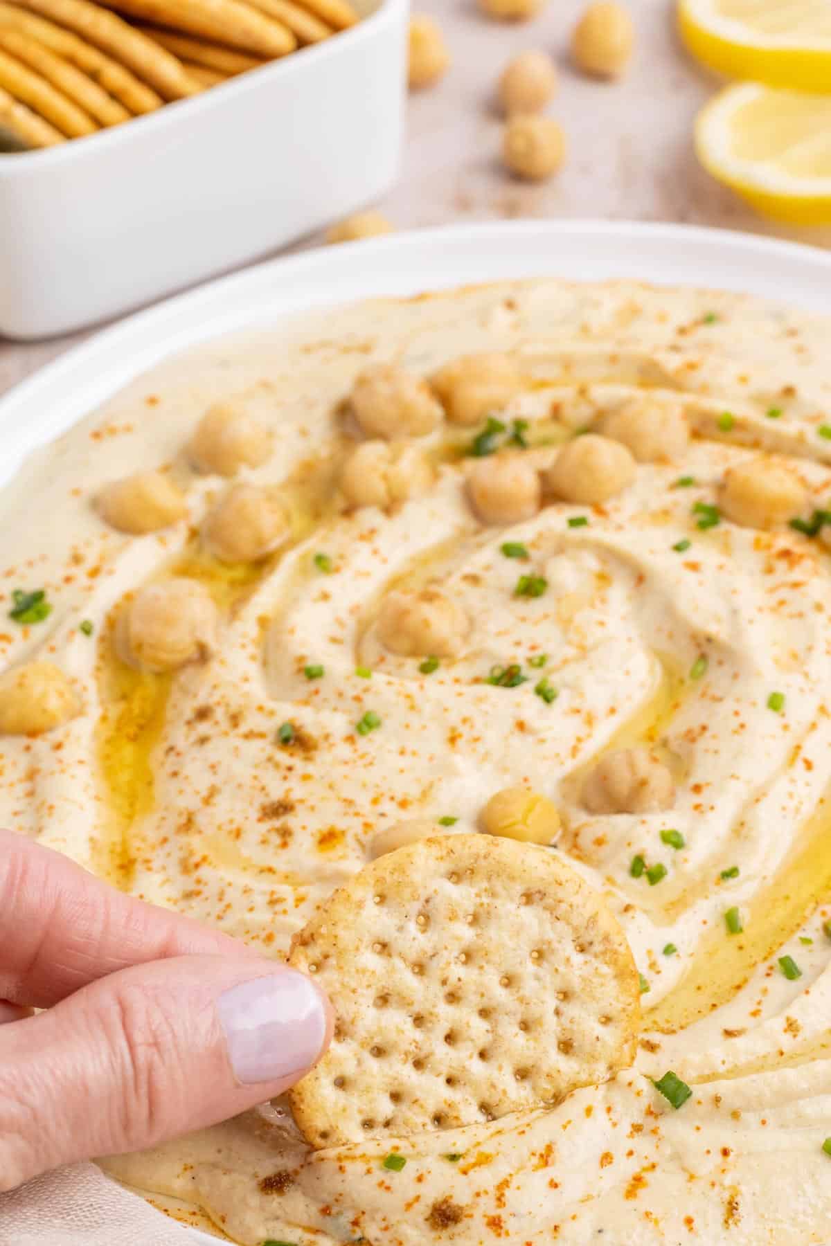 A hand scooping a cracker into hummus.