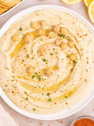 A bowl of creamy garlic hummus garnished with olive oil, red pepper, and chickpeas.
