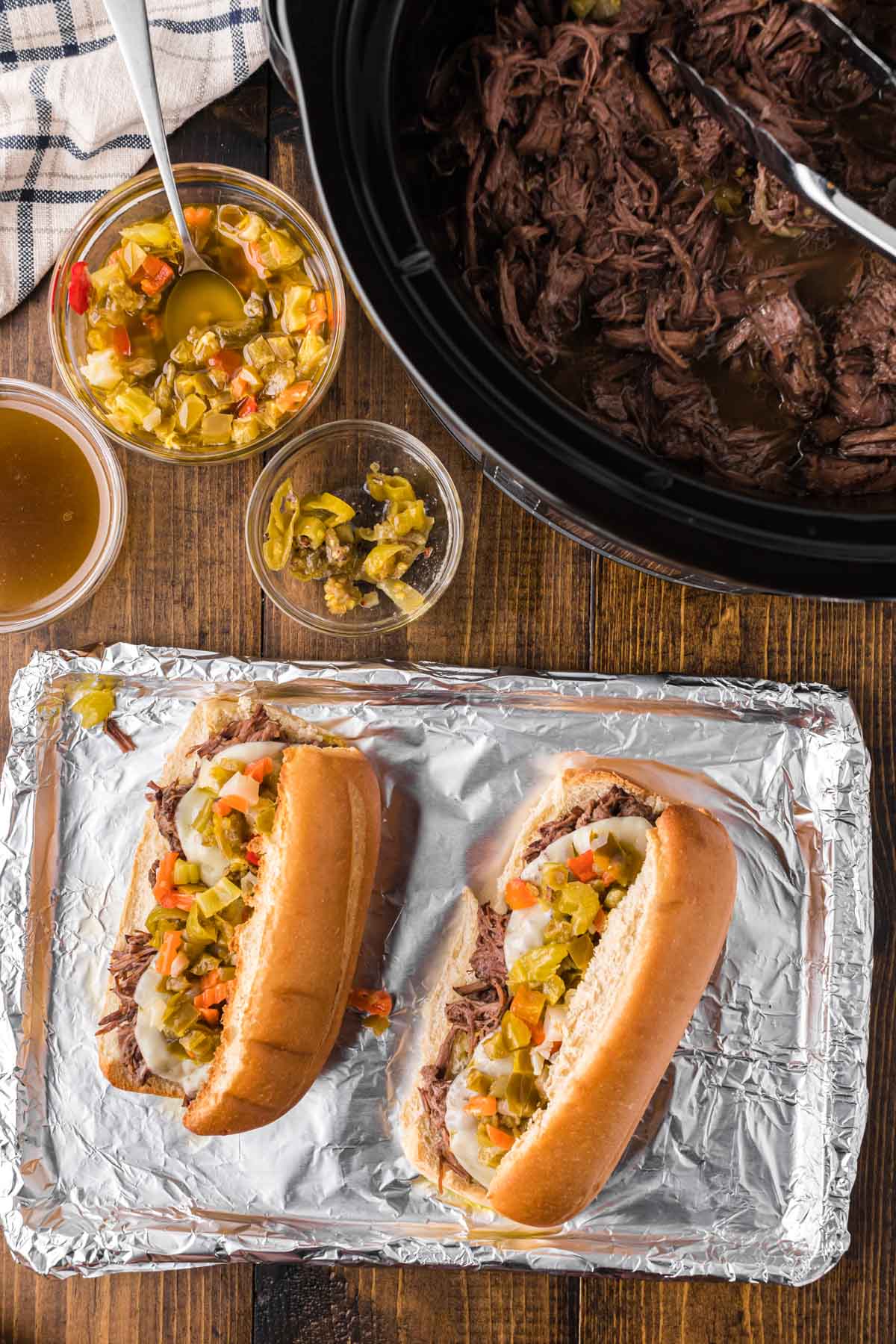 Two Italian Beef sandwiches on a baking sheet with the slow cooker full of beef set on a wooden table.