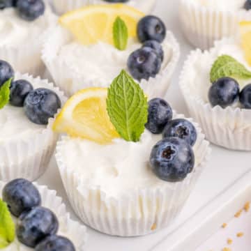 A tray of no-bake mini lemon cheesecakes garnished with lemon slice, blueberries and mint.