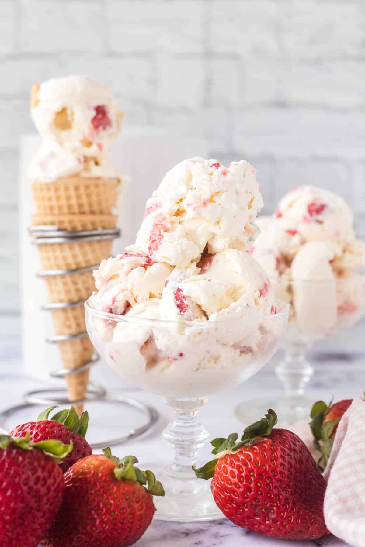 Two glass dessert bowls filled with scoops of strawberry cheesecake ice cream and a cone filled in the background.