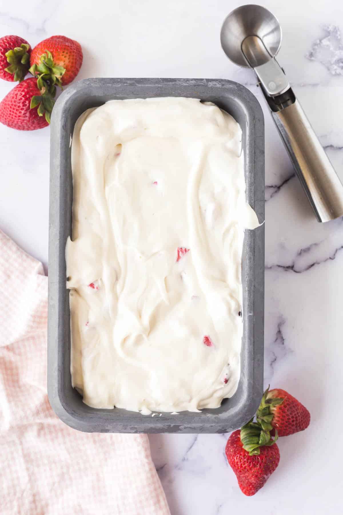 Frozen Strawberry Cheesecake Ice Cream in a loaf pan on a marble table with an ice cream scoop.