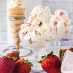 Two glass dessert bowls filled with scoops of no-churn strawberry cheesecake ice cream and a cone filled in the background.