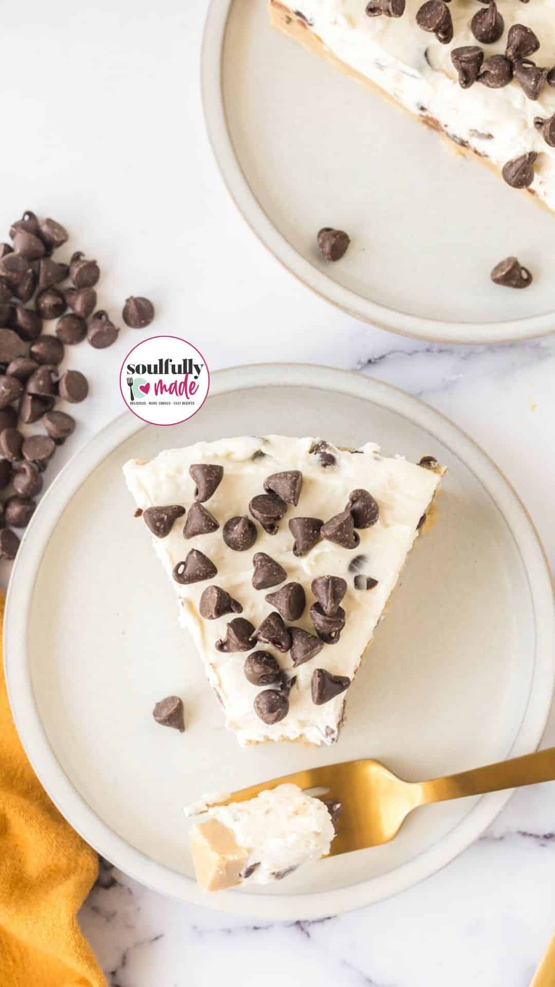 A slice of the delicious no-bake Cookie Dough Cheesecake topped with chocolate chips on a white plate. A gold fork has taken out a bite.